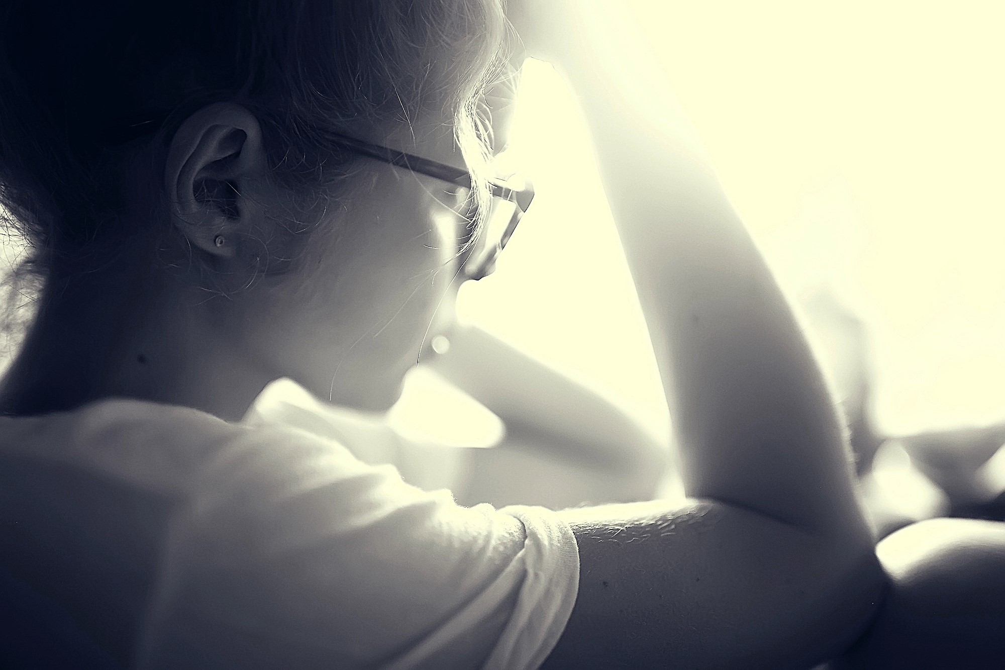 Black and white photo of a person in profile, wearing glasses and a white shirt. Their hand rests on their head, and they are looking towards a bright light, creating a soft, reflective mood.