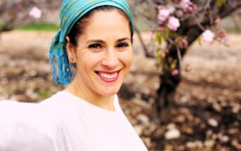 A person wearing a turquoise headscarf and a white top smiles for a selfie in an outdoor setting with blurred cherry blossoms in the background.