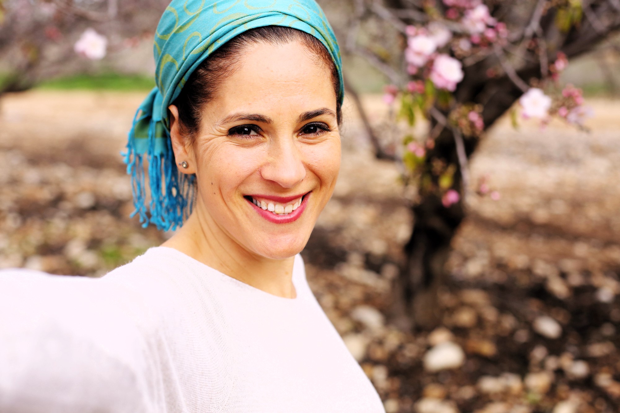 A person wearing a turquoise headscarf and a white top smiles for a selfie in an outdoor setting with blurred cherry blossoms in the background.