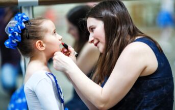 A woman is applying lipstick to a young girl dressed in a ballet outfit with a blue hair accessory. They are in front of a mirror, and the setting appears to be a dance studio. The girl looks up attentively at the woman.