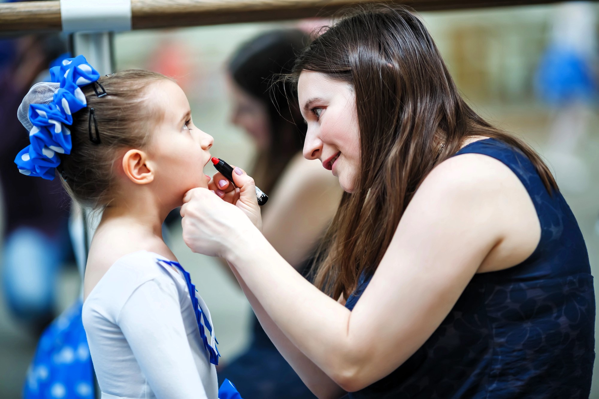 A woman is applying lipstick to a young girl dressed in a ballet outfit with a blue hair accessory. They are in front of a mirror, and the setting appears to be a dance studio. The girl looks up attentively at the woman.