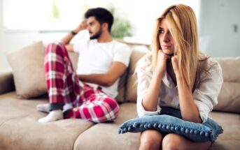 A woman with blonde hair sits on a couch, holding a cushion and looking contemplative. A man in a white shirt and red plaid pants sits behind her, looking away. Both appear to be deep in thought. The room is softly lit.