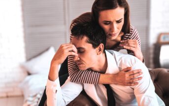 A woman gently embraces a man who is sitting with his eyes closed, appearing stressed. She wears a striped top, and he is in a white shirt with a loosened black tie. They are in a softly lit room with blurred furniture in the background.