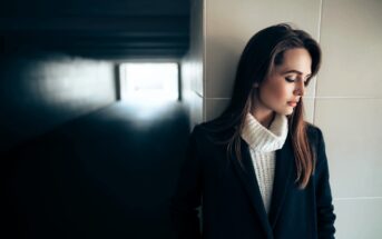 A woman with long brown hair leans against a tiled wall, looking contemplative. She wears a dark coat over a white turtleneck sweater. Sunlight shines through an opening at the end of a shadowy corridor behind her.