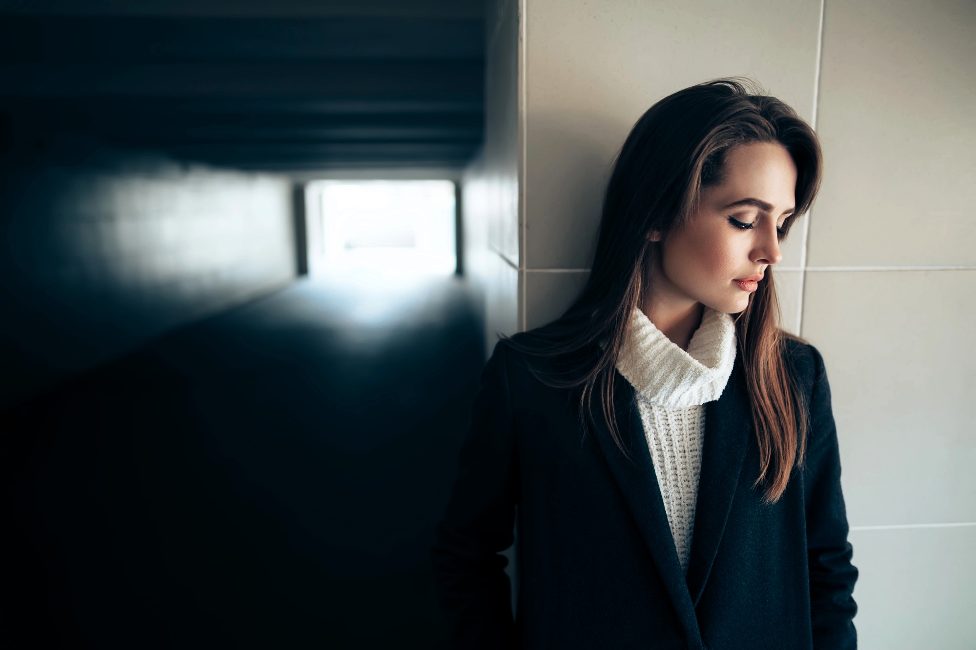 A woman with long brown hair leans against a tiled wall, looking contemplative. She wears a dark coat over a white turtleneck sweater. Sunlight shines through an opening at the end of a shadowy corridor behind her.