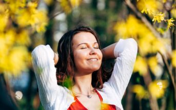 A woman stands outdoors with her eyes closed, smiling and enjoying the sunshine. She wears a white sweater over a red top and holds her hands behind her head. Yellow flowers and greenery surround her, creating a serene and cheerful atmosphere.