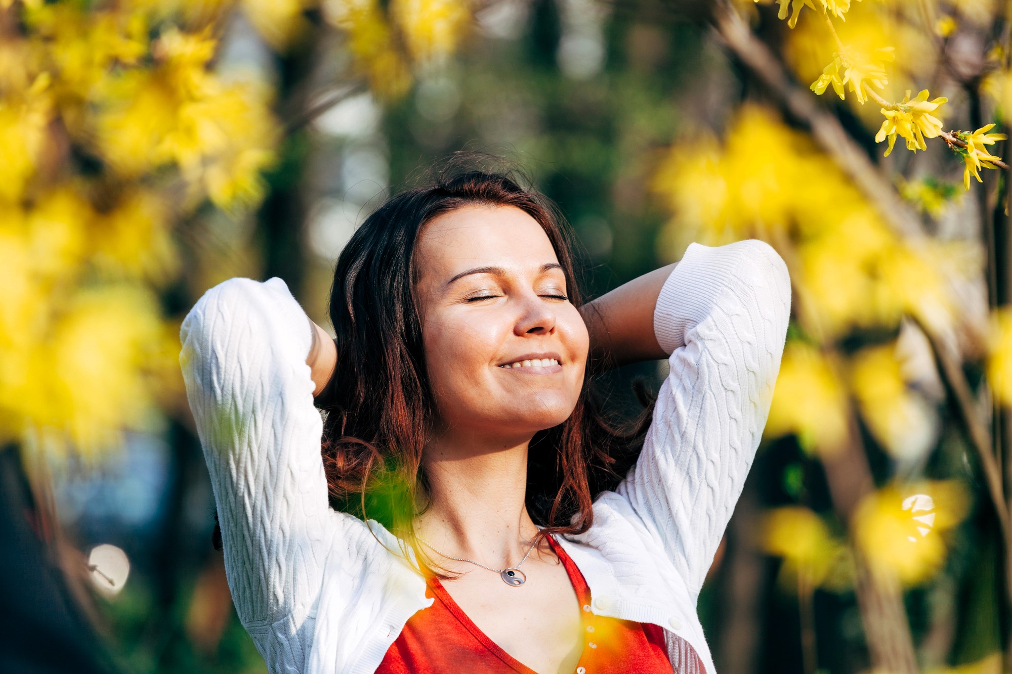 A woman stands outdoors with her eyes closed, smiling and enjoying the sunshine. She wears a white sweater over a red top and holds her hands behind her head. Yellow flowers and greenery surround her, creating a serene and cheerful atmosphere.