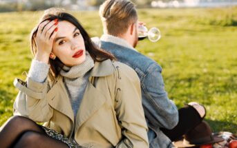 A woman in a beige coat sits on grass, looking at the camera with her hand in her hair. She has red lipstick and painted nails. A man, facing away, drinks from a glass. They are outdoors on a sunny day with blurry greenery in the background.