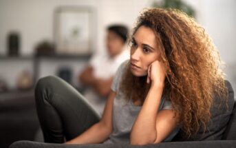 A woman with curly hair, wearing a gray t-shirt, sits on a couch looking pensive. Her elbow rests on her knee and her chin on her hand. The background is blurred, with a person sitting in another area of the room.