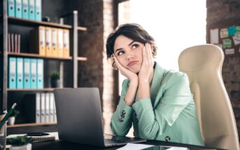 A woman in a light green blazer sits at a desk in front of a laptop, resting her chin on her hands and looking contemplative. The office setting includes shelves with colorful binders and a window letting in natural light.