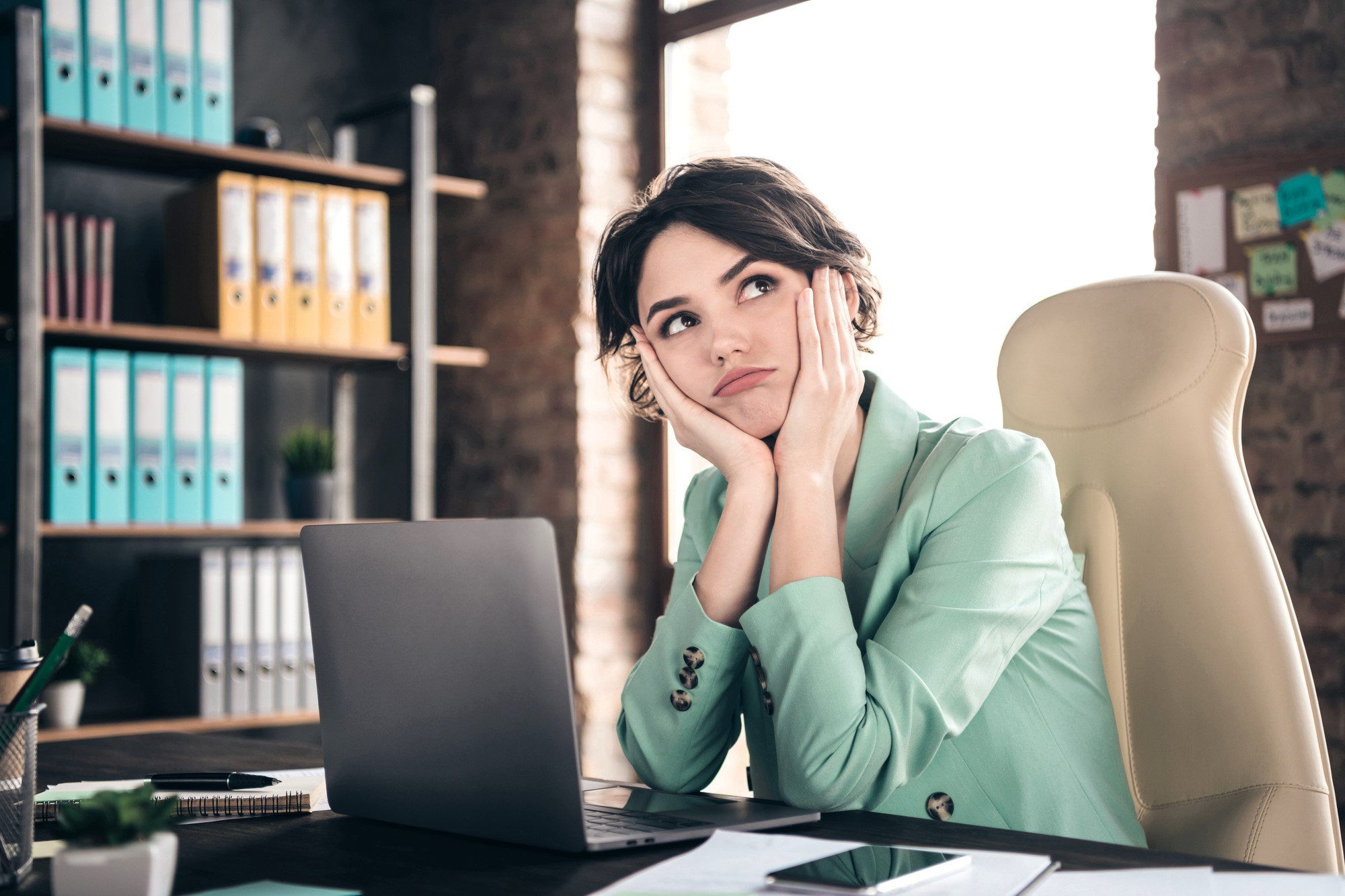 A woman in a light green blazer sits at a desk in front of a laptop, resting her chin on her hands and looking contemplative. The office setting includes shelves with colorful binders and a window letting in natural light.