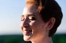 A person with short red hair and freckles smiles with eyes closed, facing the sun. They are wearing dangle earrings. The background is blurred, suggesting an outdoor setting with a clear sky.