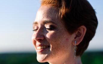 A person with short red hair and freckles smiles with eyes closed, facing the sun. They are wearing dangle earrings. The background is blurred, suggesting an outdoor setting with a clear sky.