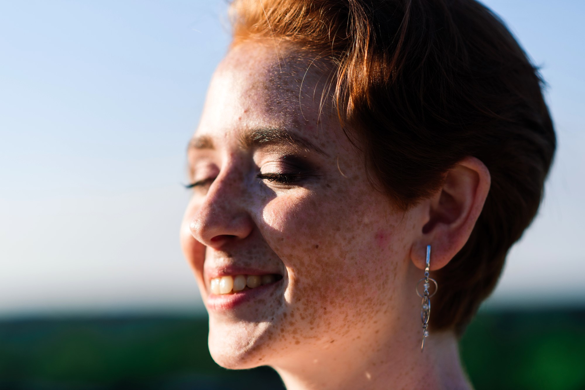 A person with short red hair and freckles smiles with eyes closed, facing the sun. They are wearing dangle earrings. The background is blurred, suggesting an outdoor setting with a clear sky.