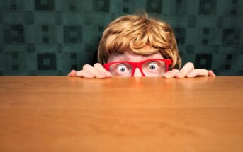 A person with short blonde hair and red glasses peeks nervously over a wooden table. Eyes wide open, they grip the table's edge. The background is a dark green patterned curtain.