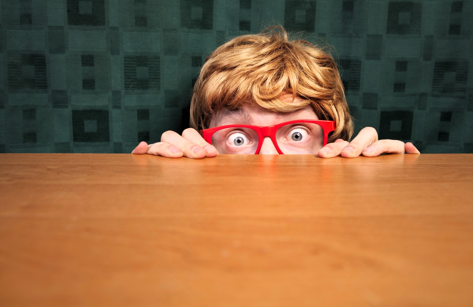 A person with short blonde hair and red glasses peeks nervously over a wooden table. Eyes wide open, they grip the table's edge. The background is a dark green patterned curtain.