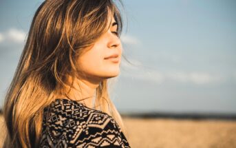 A woman with long hair stands in a field under a blue sky, facing sideways with eyes closed. She wears a patterned top and appears serene, with the wind gently blowing her hair.