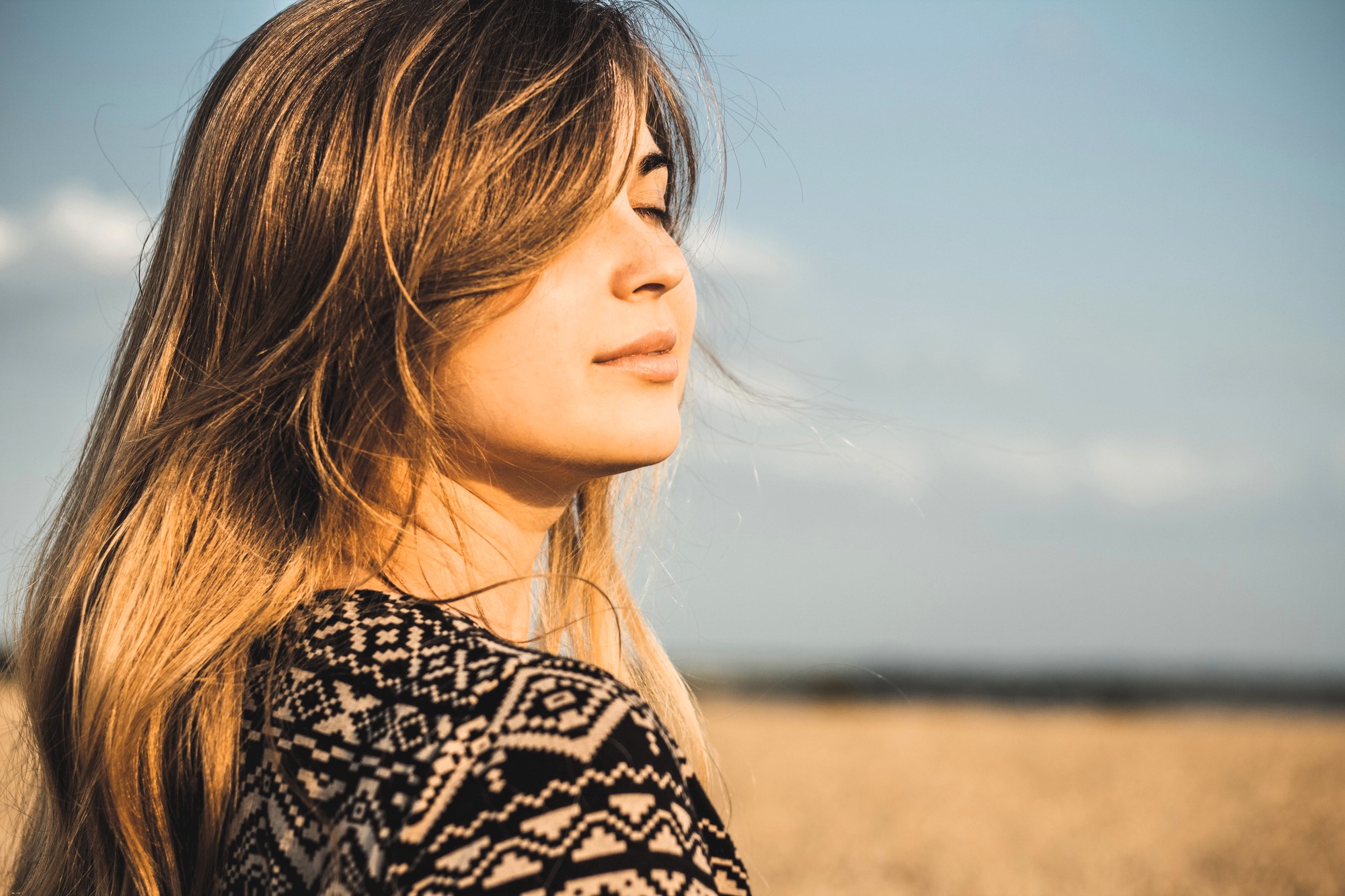 A woman with long hair stands in a field under a blue sky, facing sideways with eyes closed. She wears a patterned top and appears serene, with the wind gently blowing her hair.