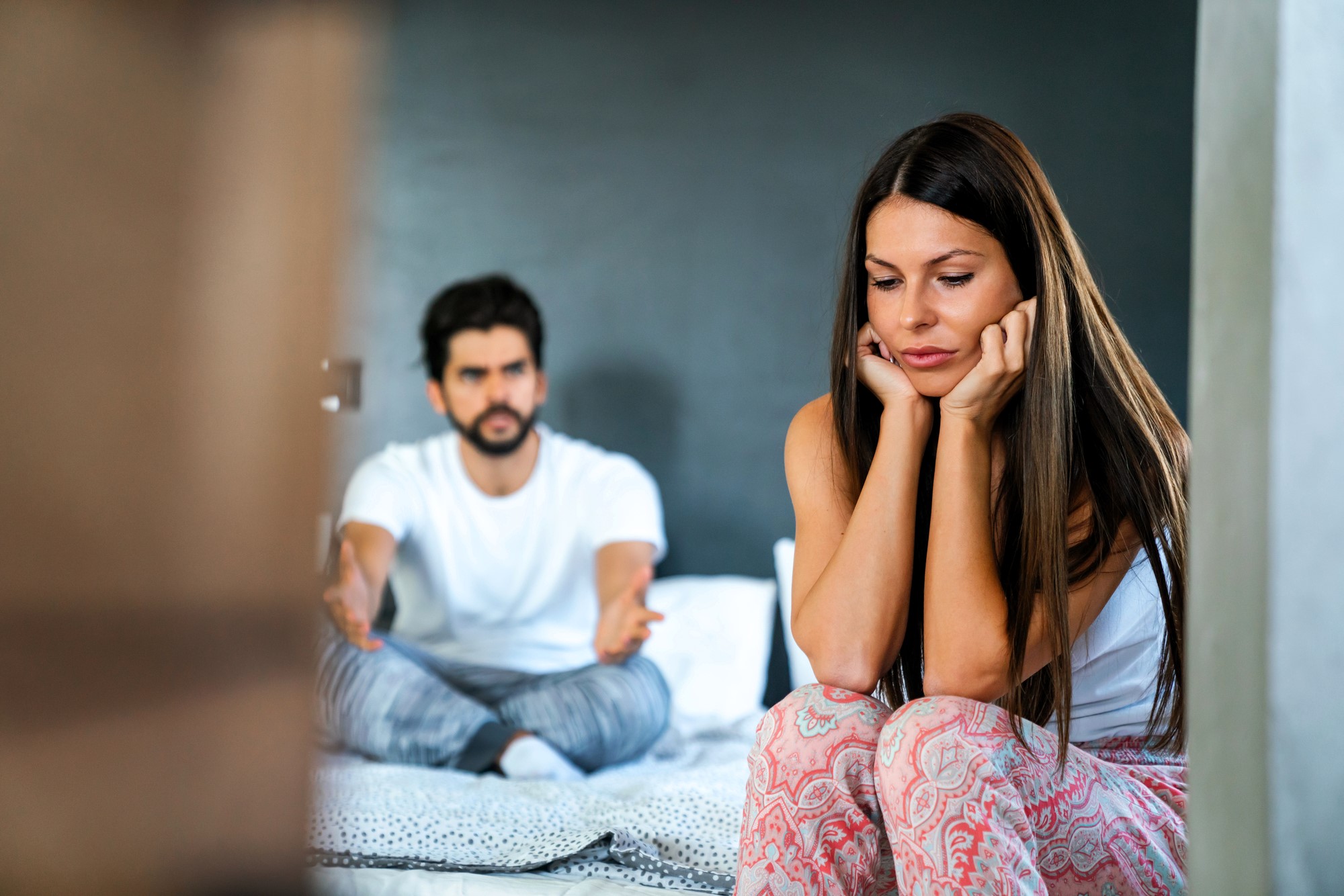 A woman sits on the edge of a bed, looking down with a pensive expression, her hands on her cheeks. A man, seated on the bed behind her, gestures with a concerned expression. Both wear casual clothing.