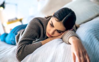 A woman lies on a bed, leaning on her right arm, with a thoughtful expression. She is wearing a gray sweater and blue jeans. The background is softly blurred, suggesting a cozy indoor setting.