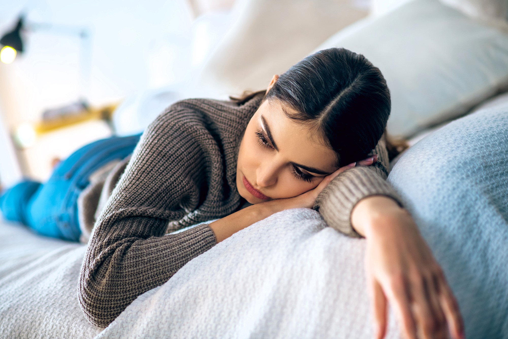 A woman lies on a bed, leaning on her right arm, with a thoughtful expression. She is wearing a gray sweater and blue jeans. The background is softly blurred, suggesting a cozy indoor setting.