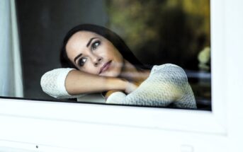 A woman with long dark hair leans on a window, looking outside with a thoughtful expression. She rests her chin on her crossed arms and wears a white knit sweater. The background is softly blurred.