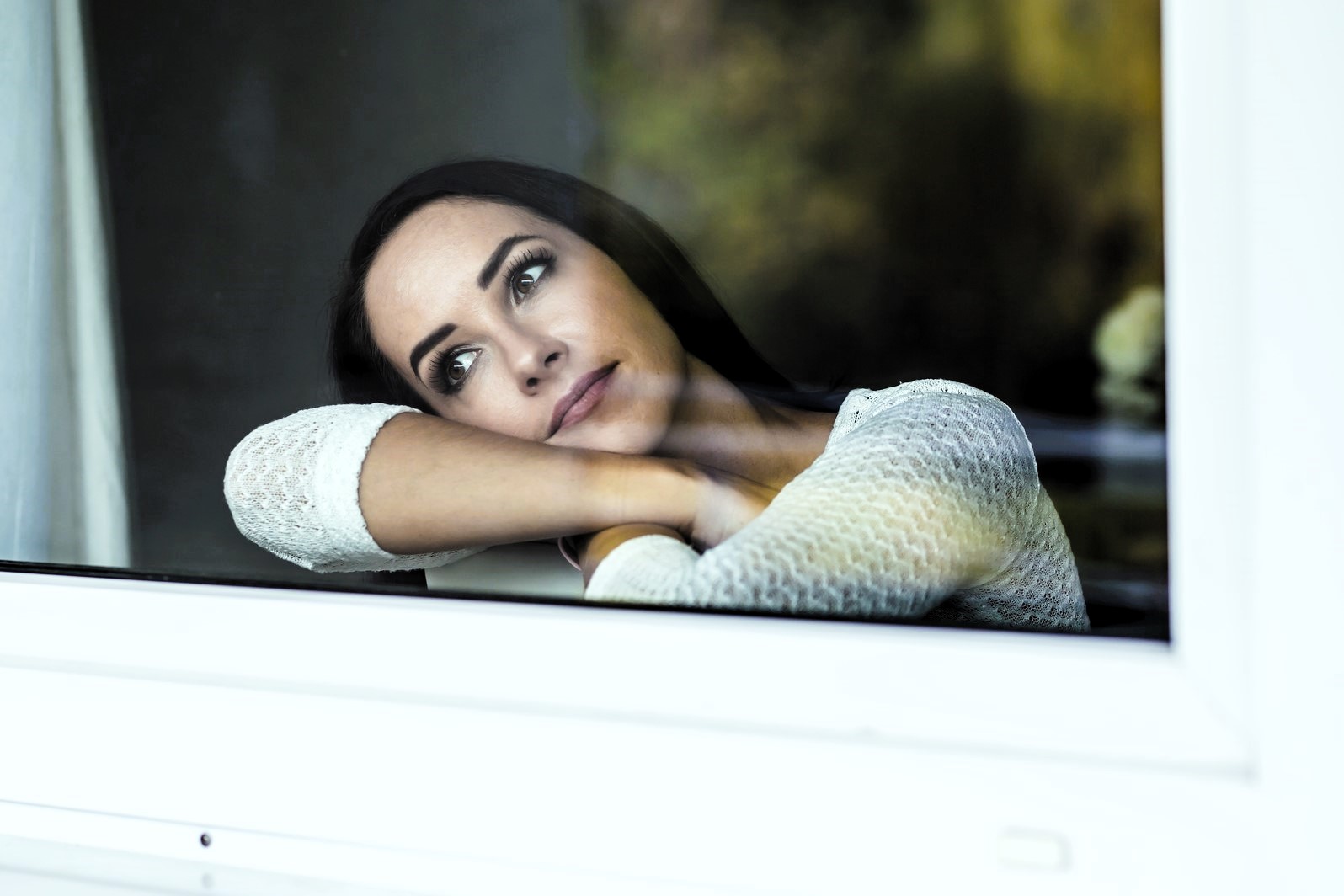 A woman with long dark hair leans on a window, looking outside with a thoughtful expression. She rests her chin on her crossed arms and wears a white knit sweater. The background is softly blurred.