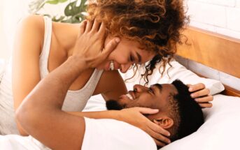 A couple smiling affectionately at each other while lying on a bed. The woman is on top, gently holding the man's face. Both are looking into each other's eyes. A plant is in the background.