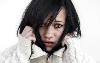 A person with black hair, wearing a white cable-knit sweater, covers part of their face with the sweater's collar. They have an intense expression, with hair partially falling over their eyes, against a plain white background.