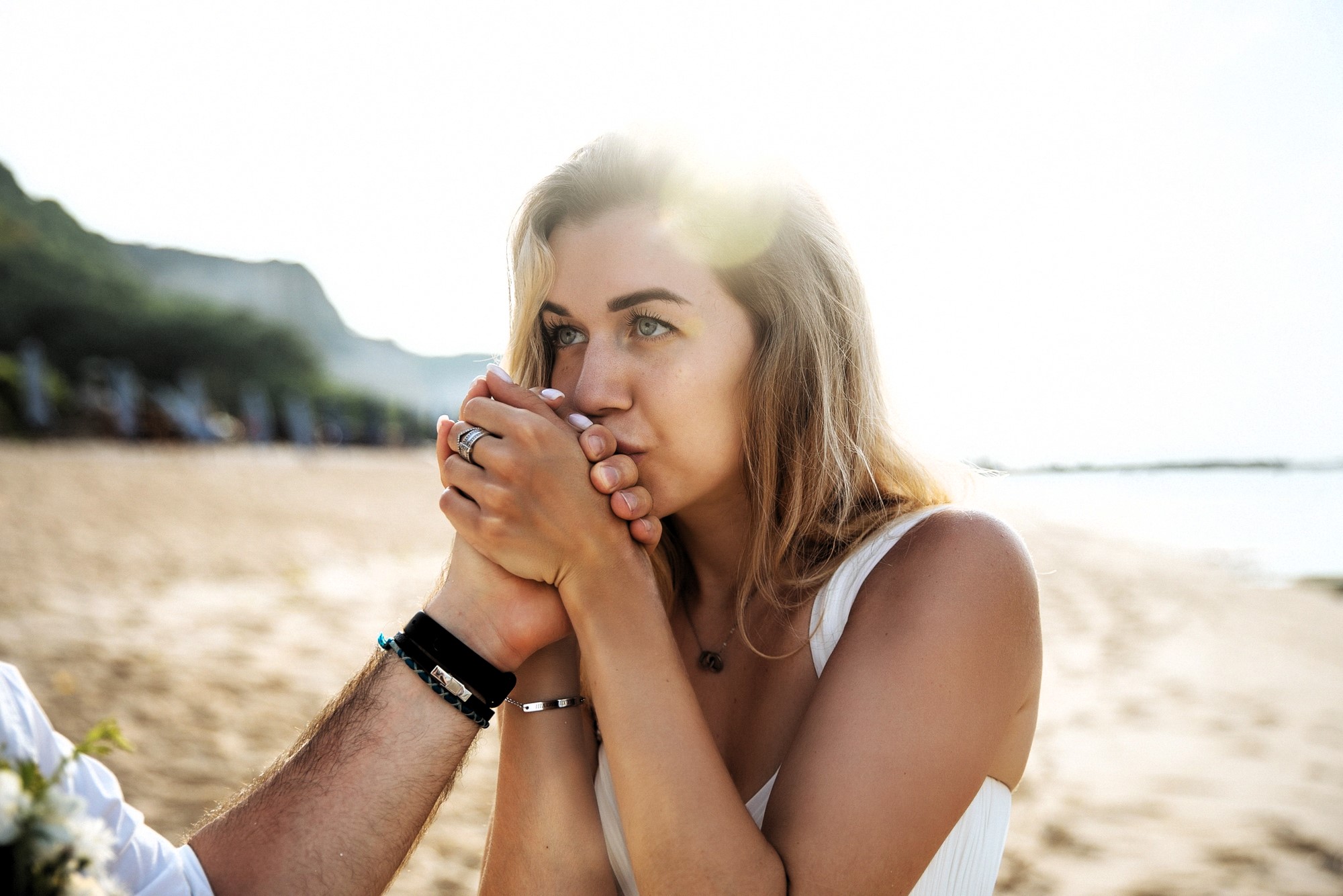 A woman with long hair holds and kisses a person's hand while sitting on a sandy beach. She is wearing a white sleeveless top. The background features a sunny beach scene with greenery and blurred silhouettes of people.