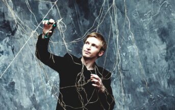 A man in a black suit holds green scissors and looks up while tangled in a web of strings against a textured gray background. He appears focused as he attempts to cut the strings above him.