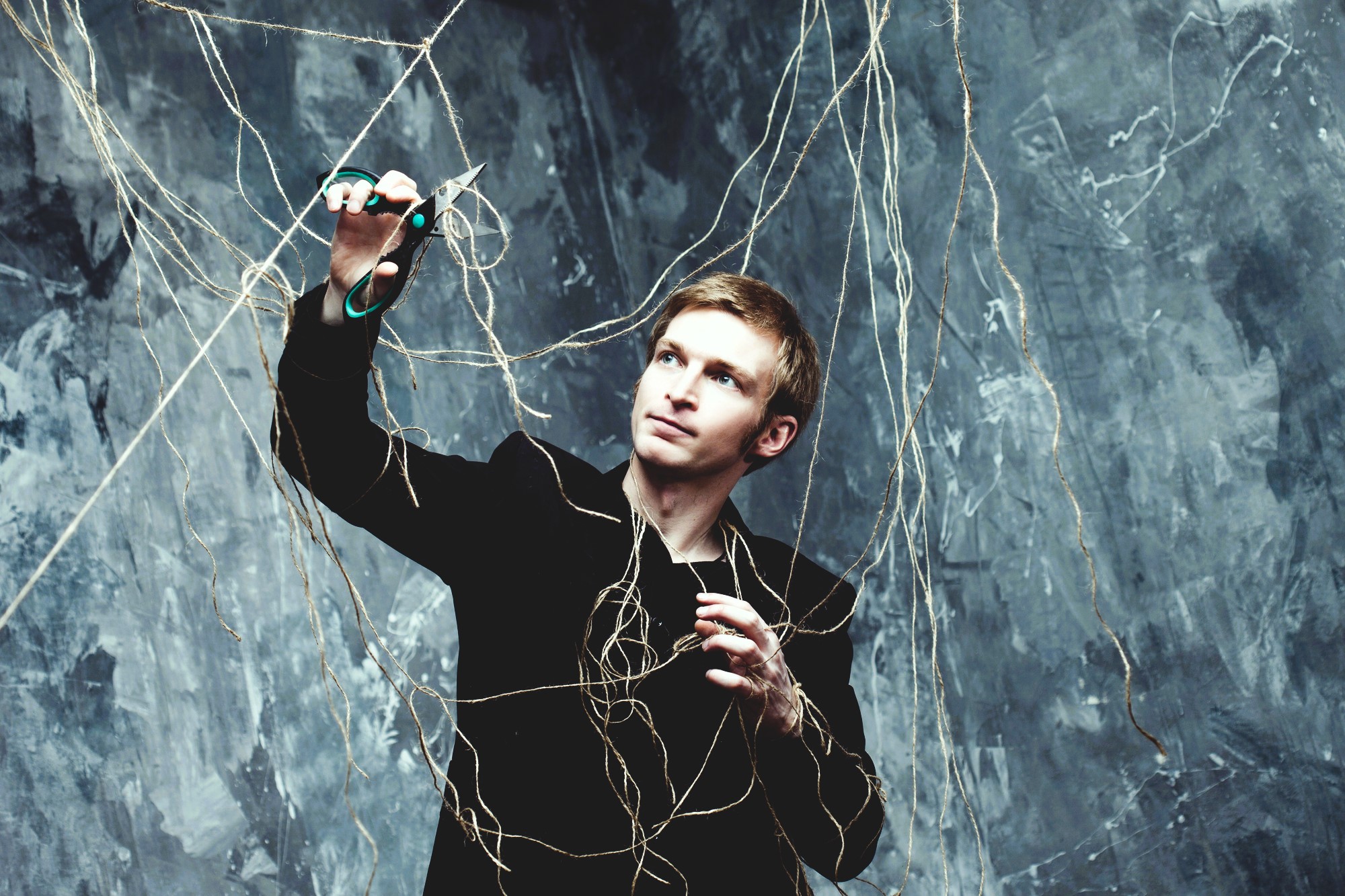 A man in a black suit holds green scissors and looks up while tangled in a web of strings against a textured gray background. He appears focused as he attempts to cut the strings above him.