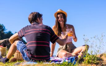 A man and a woman sitting on a blanket outdoors, toasting with plastic cups. The woman is wearing a straw hat and a white top, smiling under a clear blue sky. They are surrounded by grass and small plants, with a picnic basket nearby.