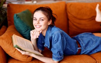 Woman in a denim outfit lying on an orange sofa, smiling, and holding an open book. An emerald green pillow is beside her. The background is softly blurred, suggesting an indoor setting.