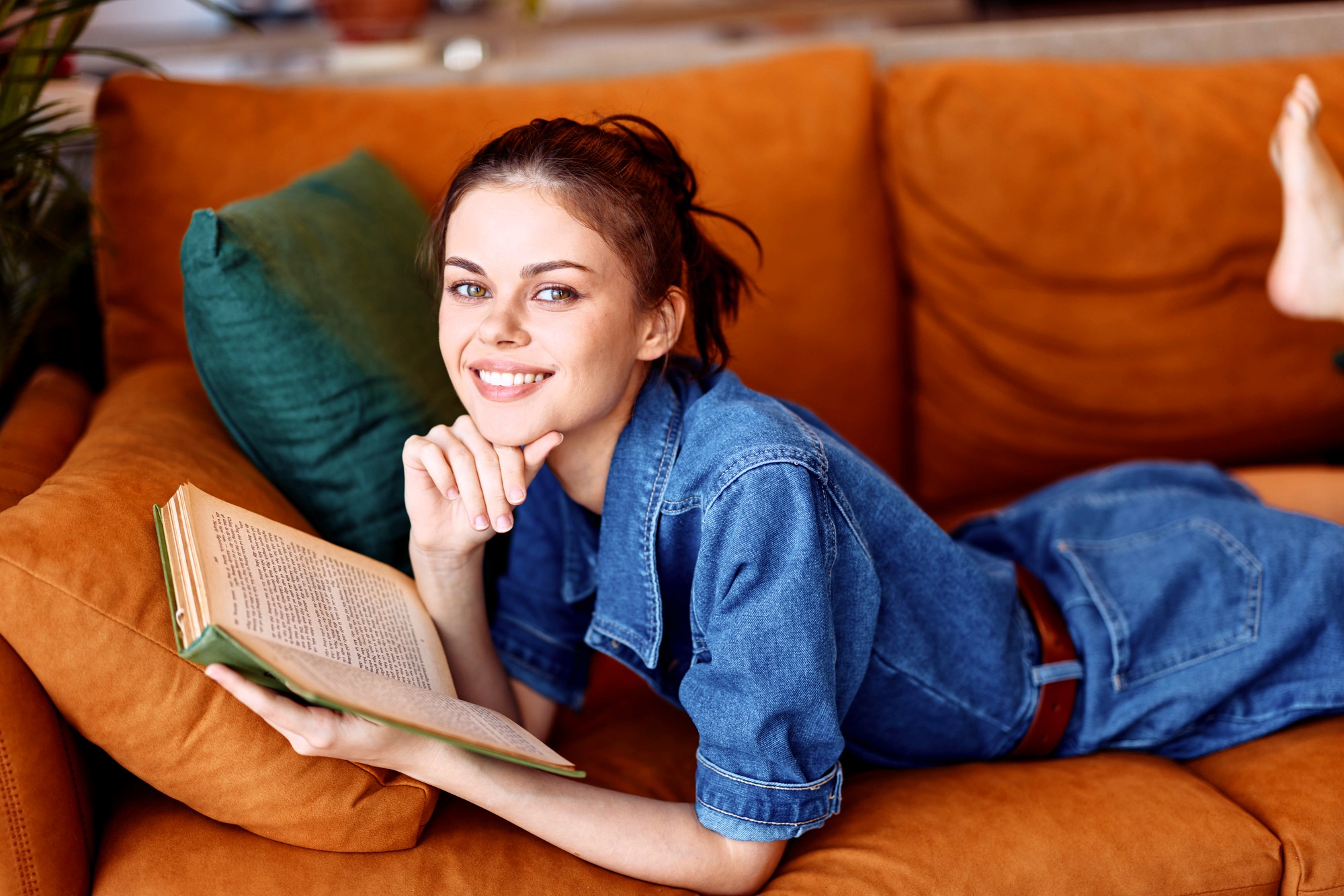 Woman in a denim outfit lying on an orange sofa, smiling, and holding an open book. An emerald green pillow is beside her. The background is softly blurred, suggesting an indoor setting.