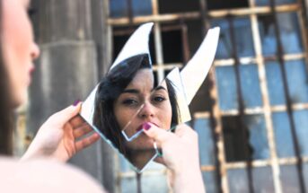 A person holds a broken mirror reflecting their face. The background features an old window with rusted bars. The focus is on the person's reflection, creating a fragmented, artistic composition.