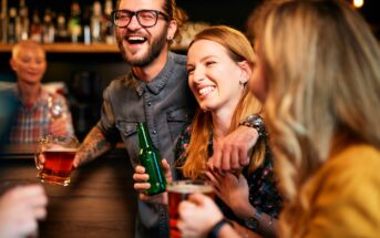 A group of friends joyfully laugh and chat at a bar. A man with glasses holds a beer mug, and a woman with a floral top clutches a green bottle. The background shows a bar with various bottles. The atmosphere is lively and warm.