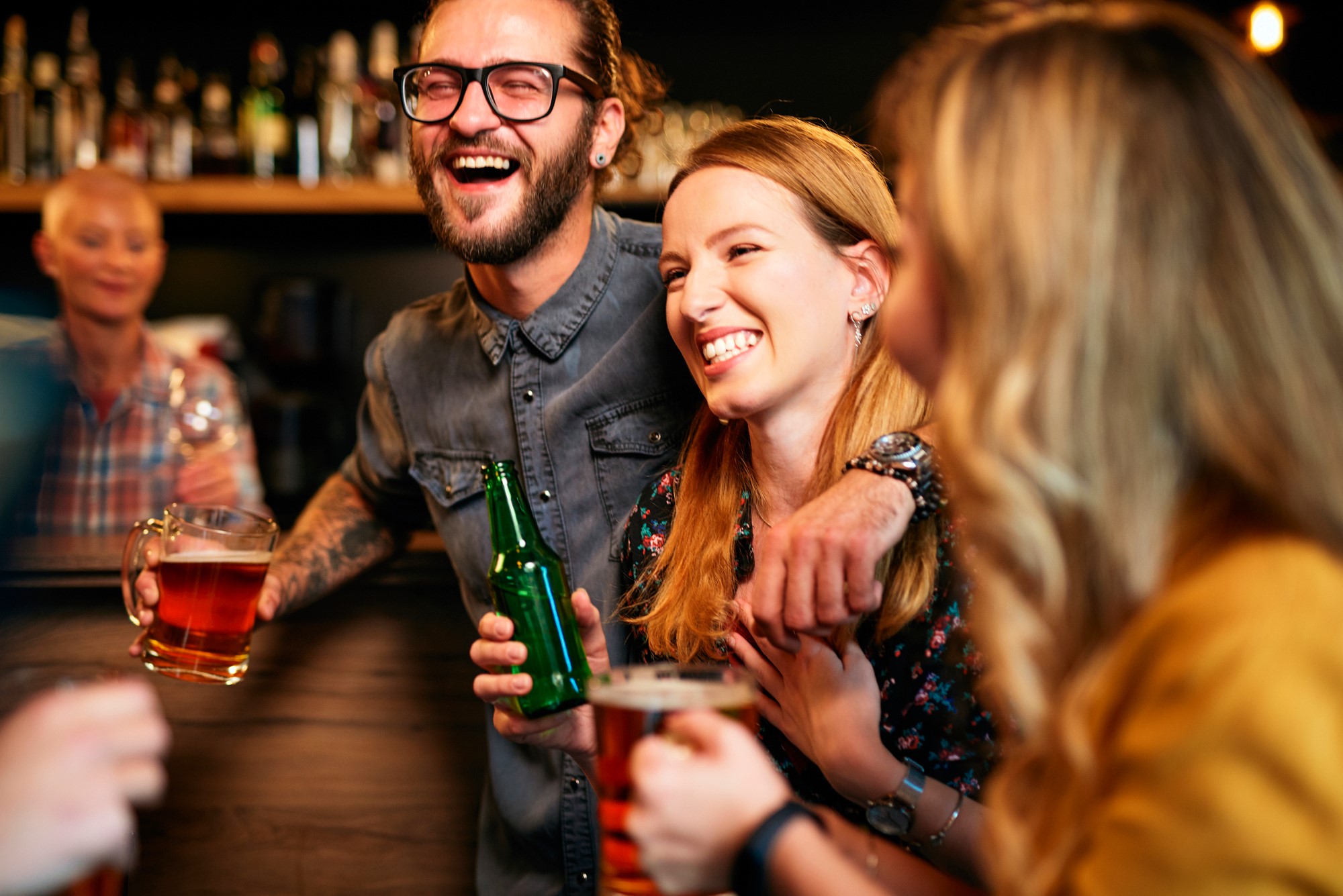 A group of friends joyfully laugh and chat at a bar. A man with glasses holds a beer mug, and a woman with a floral top clutches a green bottle. The background shows a bar with various bottles. The atmosphere is lively and warm.