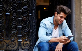 Man sitting on a step, wearing a light denim jacket and jeans, looking at his smartphone and smiling. The background features an ornate wrought iron gate.