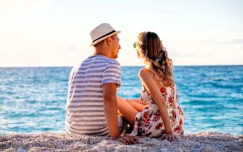 A couple sits together on a beach, facing the ocean. The man wears a striped shirt and a hat, while the woman is in a floral dress and sunglasses. The sun is setting, casting a warm glow on the water and creating a serene atmosphere.