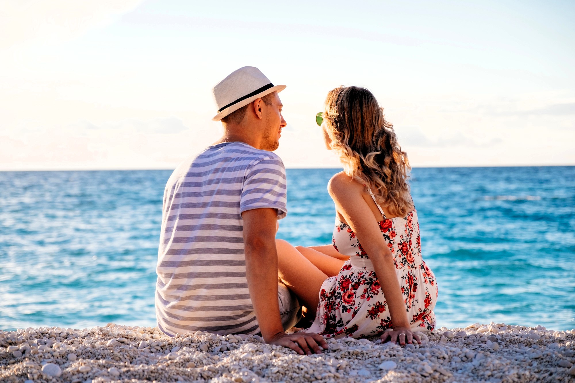 A couple sits together on a beach, facing the ocean. The man wears a striped shirt and a hat, while the woman is in a floral dress and sunglasses. The sun is setting, casting a warm glow on the water and creating a serene atmosphere.