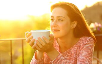 A woman is sitting outdoors in warm sunlight, holding a large white mug with both hands. She has long brown hair and is wearing a pink striped top. She looks content as she gazes into the distance.