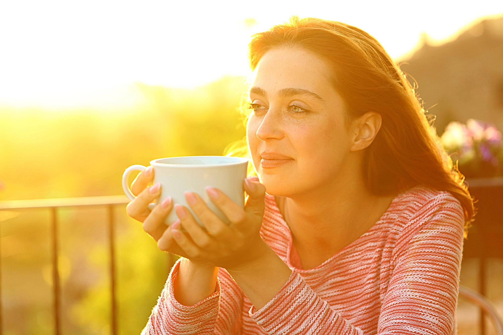 A woman is sitting outdoors in warm sunlight, holding a large white mug with both hands. She has long brown hair and is wearing a pink striped top. She looks content as she gazes into the distance.