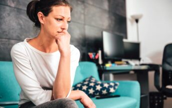 Woman in a white shirt sits thoughtfully on a teal sofa with a star-patterned pillow. She rests her chin on her hand, looking contemplative. In the background, a desk with a computer and a chair is visible.