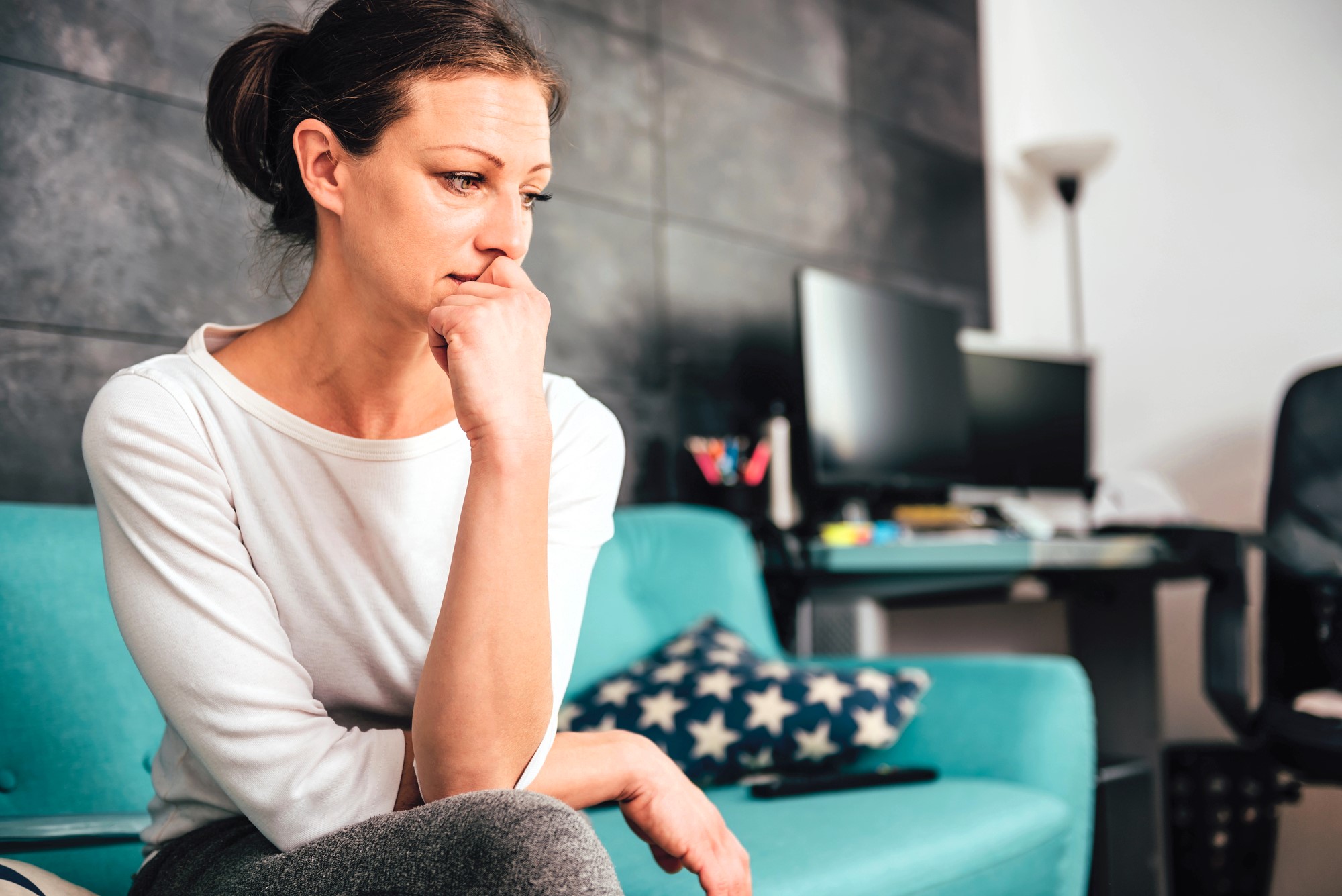 Woman in a white shirt sits thoughtfully on a teal sofa with a star-patterned pillow. She rests her chin on her hand, looking contemplative. In the background, a desk with a computer and a chair is visible.