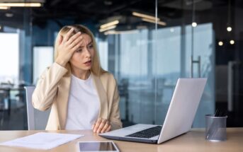 A woman with long blonde hair sits at a desk in an office, looking at a laptop with a concerned expression. She holds her forehead with one hand. Papers and a tablet are on the desk. She wears a beige blazer and white shirt.