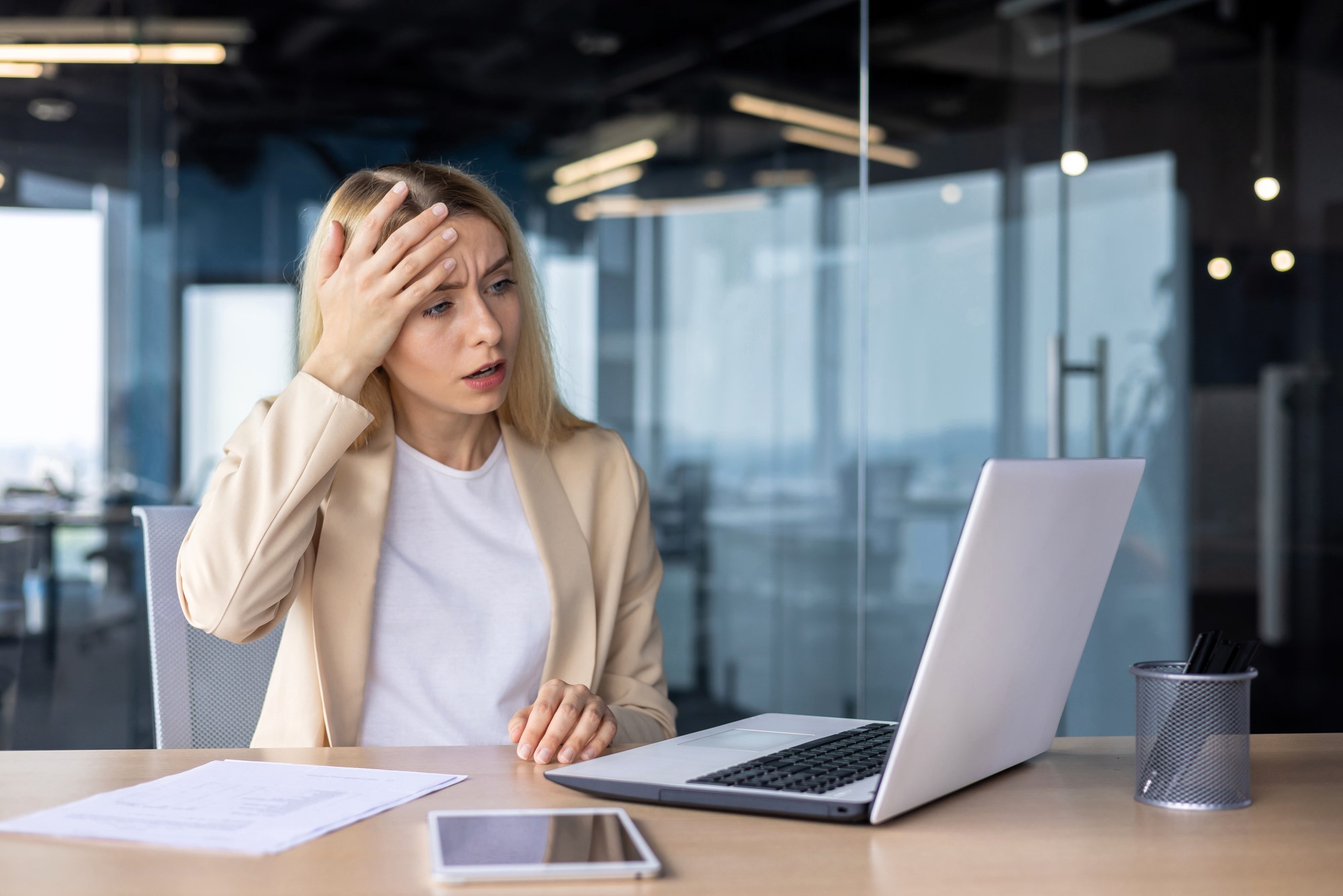 A woman with long blonde hair sits at a desk in an office, looking at a laptop with a concerned expression. She holds her forehead with one hand. Papers and a tablet are on the desk. She wears a beige blazer and white shirt.