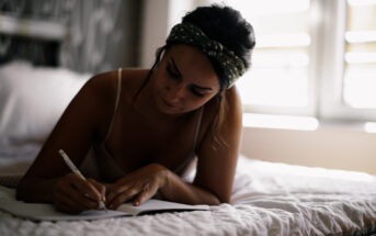 A woman with a headband is lying on her stomach on a bed, focused on writing in a notebook. She is in a softly lit room with a large window in the background.