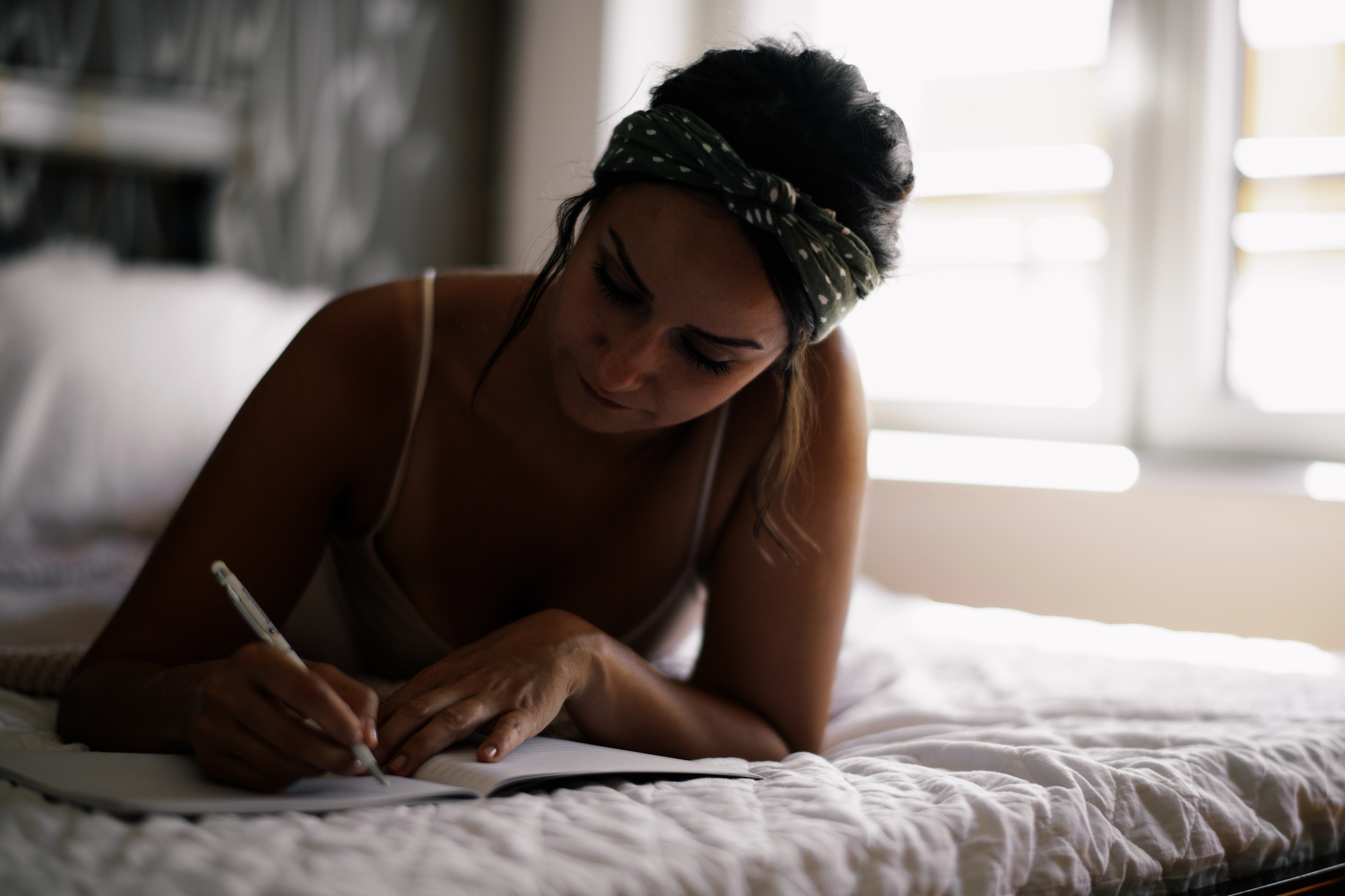 A woman with a headband is lying on her stomach on a bed, focused on writing in a notebook. She is in a softly lit room with a large window in the background.