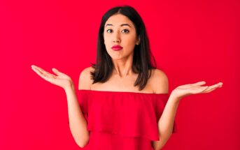 A woman with long dark hair is wearing a red off-the-shoulder top against a red background. She has an expression of confusion or uncertainty, with her arms outstretched and palms facing up.